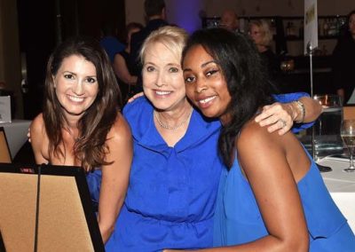 Three women smiling at a dinner event, two in matching blue dresses.