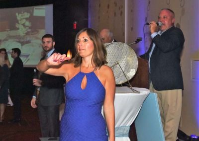 A woman in a blue dress playing a game at an event, pointing a dart forward, with a man speaking into a microphone in the background.