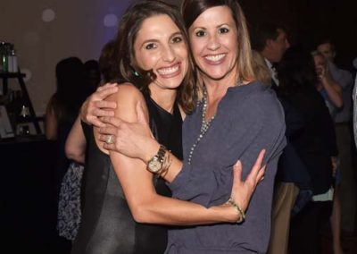 Two women smiling and embracing at a social event with dim lighting and a blurred background.