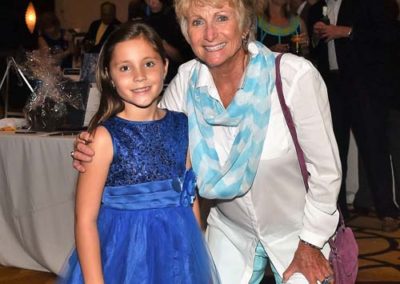 A young girl in a blue dress and an older woman in white pants and a blue scarf smiling together at a social event.
