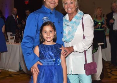 A chef in a blue jacket poses with an elderly woman and a young girl in a blue dress at an indoor event.