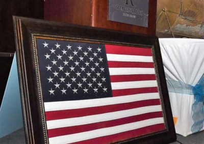 An american flag displayed in a dark ornate frame on a table, with other decorative items and a plaque in the background.