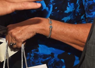 Close-up of an elderly woman's hands with a silver bracelet, holding a small bag, with a blue floral dress in the background.