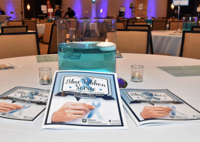 Event table with promotional materials for "the blue ribbon soiree," a large glass centerpiece with a blue liquid, and candle lighting.