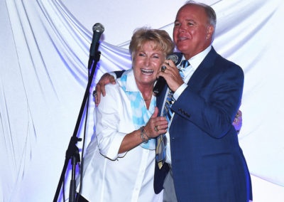 A joyful elderly couple laughing together while the man holds a microphone, standing on a stage with a white draped background.