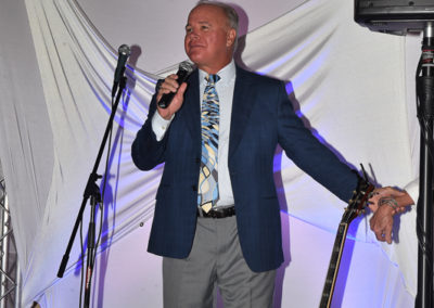 Man in a blue suit and patterned tie speaking into a microphone at an event, standing next to an acoustic guitar leaning against a draped fabric background.