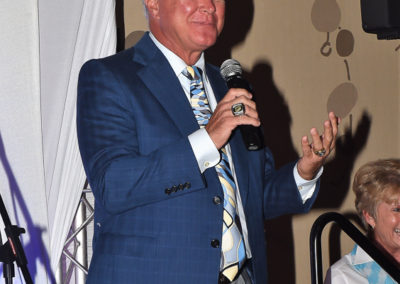 A man in a blue suit and patterned tie speaks into a microphone while gesturing with his hand at an event.
