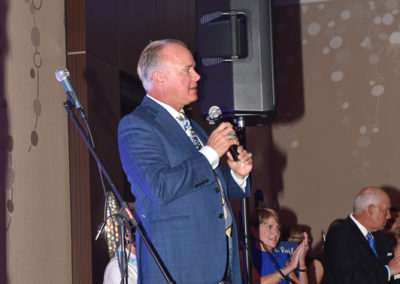 A man in a suit speaking into a microphone at a formal event with a band setting up in the background.