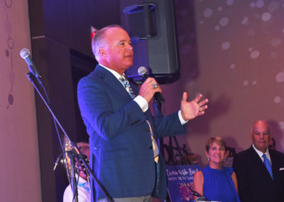 A man speaking at a microphone in a banquet hall with people watching in the background, event lights illuminate the scene.