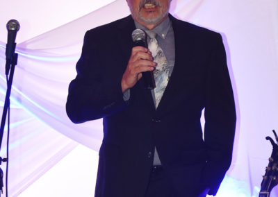 A man with a beard speaking into a microphone at an event, wearing a dark suit and a tie with a galaxy print.