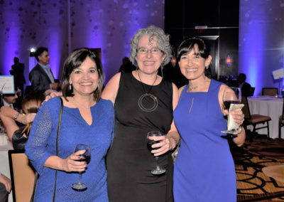 Three women in cocktail dresses, smiling with wine glasses in hand at a formal event with a blue-lit background.