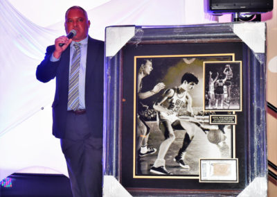 Man in a suit speaking into a microphone next to a large framed sports photograph at an event.