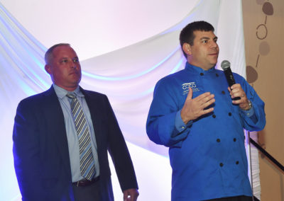 Two men on stage at an event; one in a suit and the other in a blue chef's uniform, speaking into a microphone.