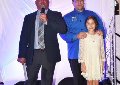 Man holding a microphone standing next to another man in a chef's uniform and a young girl in a white dress, all posing on a stage with lighting and draped background.