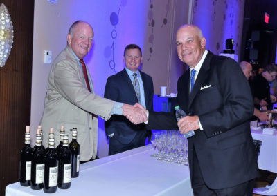 Two senior men shaking hands at an event, with another man smiling in the background and a table with wine bottles and glasses nearby.