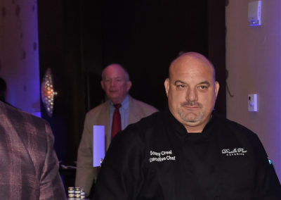 Bald chef in a black chef coat labeled "sonny creek" at a dimly lit event, with people in the background.