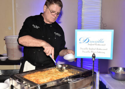 A chef serving food from a large dish at a catering event, with a sign advertising drusilla seafood restaurant.