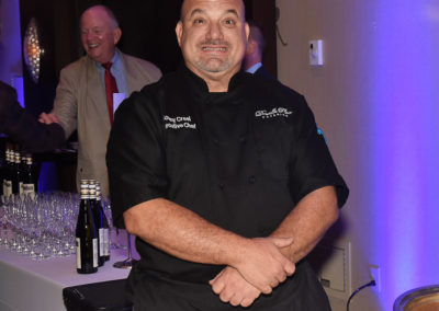 Chef in black uniform smiling with arms crossed at a catering event, with guests and a bar in the background.