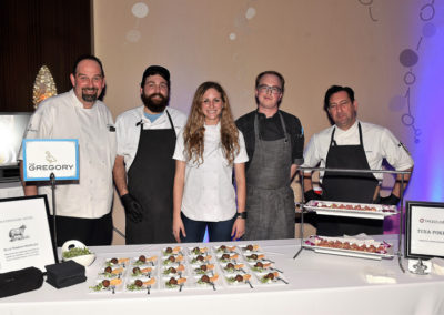 Five chefs standing behind a food tasting table with appetizers at a culinary event.