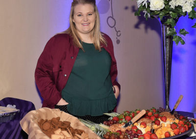 Woman standing beside a buffet table with assorted fruits, cheeses, and crackers at an event.