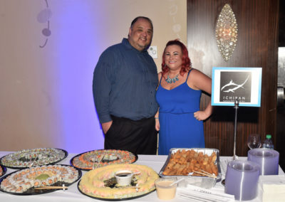 A man and a woman smiling beside a table with assorted sushi platters and appetizers at an event.