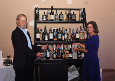 A man and a woman in formal attire selecting wine from a well-stocked bar shelf at an event.