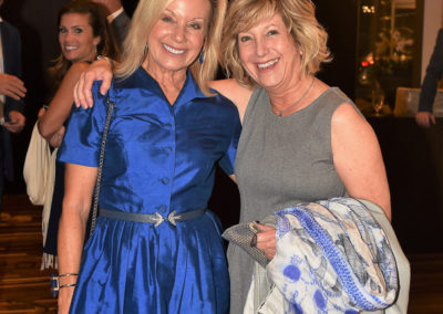 Two smiling women in elegant dresses, one in blue and the other in gray, standing arm-in-arm at a formal event.
