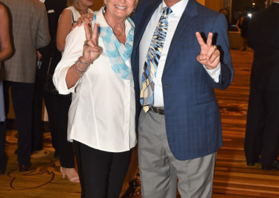 Two older adults smiling at a formal event, the woman showing a peace sign and the man in a suit and tie.