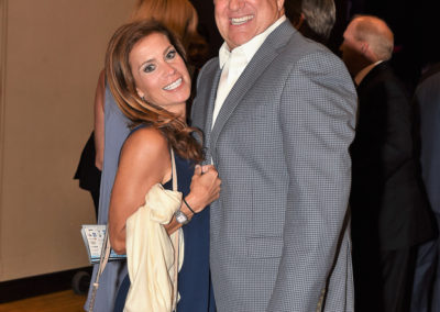 A man and a woman smiling and embracing at a formal event, the man in a gray checkered blazer and the woman in a light blue dress.