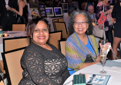 Two women smiling at a table during a formal event, with drinks and event programs in front of them.