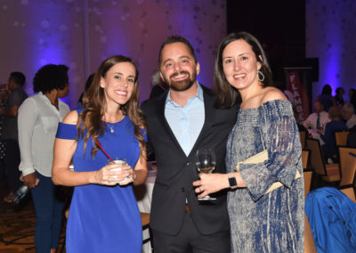 Three smiling adults, two women and one man, holding wine glasses at a social gala event with a festive backdrop.