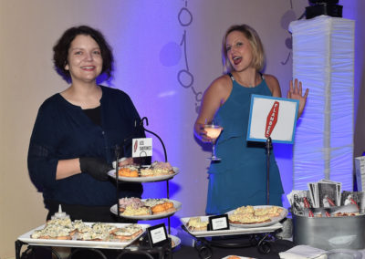 Two women standing at a buffet table with seafood dishes, one holding a sign that says "best new business," smiling and interacting at an event.