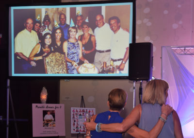 Two women facing a projector screen displaying a photo of a cheerful group celebration at a party.