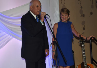 A man speaks into a microphone at an event while a smiling woman stands beside him with a guitar in the background.