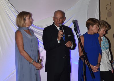 A man speaks into a microphone on stage at an event, flanked by three women listening attentively.