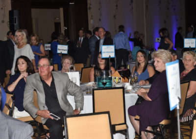 Attendees at a formal gala event, sitting at tables and conversing, with ambient lighting and event signage in the background.