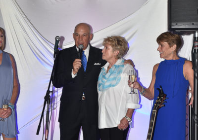 A man in a tuxedo speaks into a microphone, standing beside two women holding wine glasses at a formal event, with a draped white backdrop and a guitar nearby.