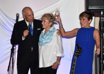 Three people speaking at a podium with a microphone, one man and two women, toasting with champagne in an event setting.