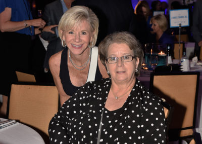 Two older women smiling at a table during an event, one with short hair wearing a polka dot blouse, the other with a pixie cut in a light blouse.