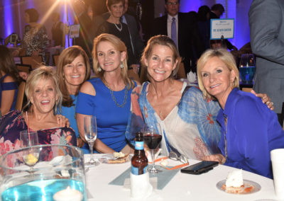 Five women smiling at a table during a gala event, with dinner plates and wine glasses in front of them.