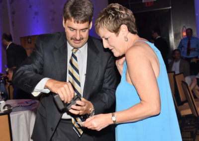 A man in a suit and a woman in a blue dress looking at a smartphone together during an event indoors.