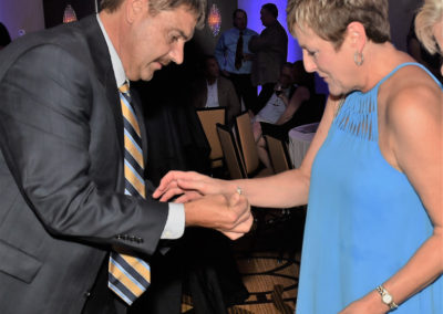A man in a suit and a woman in a blue dress dancing at an event, holding hands and looking at each other.