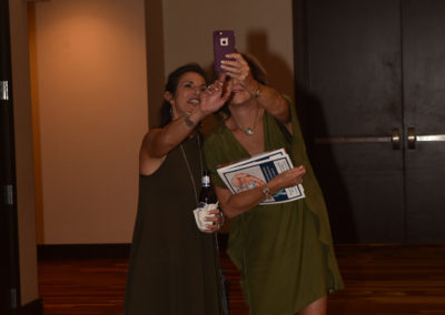 Two women taking a photograph with a smartphone in a conference hall; one poses while the other uses the phone, both dressed in green dresses.