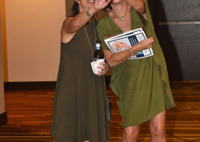 Two women in matching green dresses taking a selfie, one holding a phone and the other a booklet and drink, in a conference room.