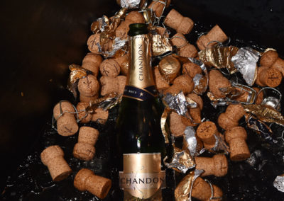 A bottle of chandon surrounded by scattered champagne corks and crumpled foil wrappers on a reflective black surface.