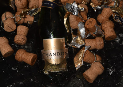 A bottle of chandon champagne surrounded by scattered corks and foil wrappers on a reflective black surface.