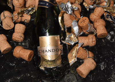 A chandon champagne bottle with its label clearly visible, surrounded by numerous corks and foil wrappers in a wet, reflective black setting.