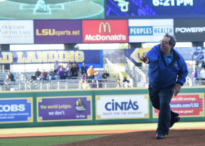 A man throws the first pitch in a baseball stadium with various advertising banners in the background.