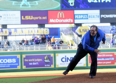A man in a blue jacket throws the first pitch at a baseball game with various advertising banners and a digital scoreboard in the background.
