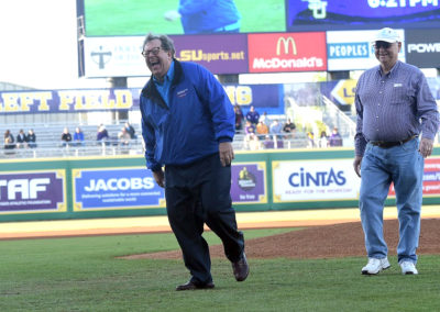 Two older men laughing on a baseball field; one in a blue jacket is playfully running, and the other in a checked shirt watches.